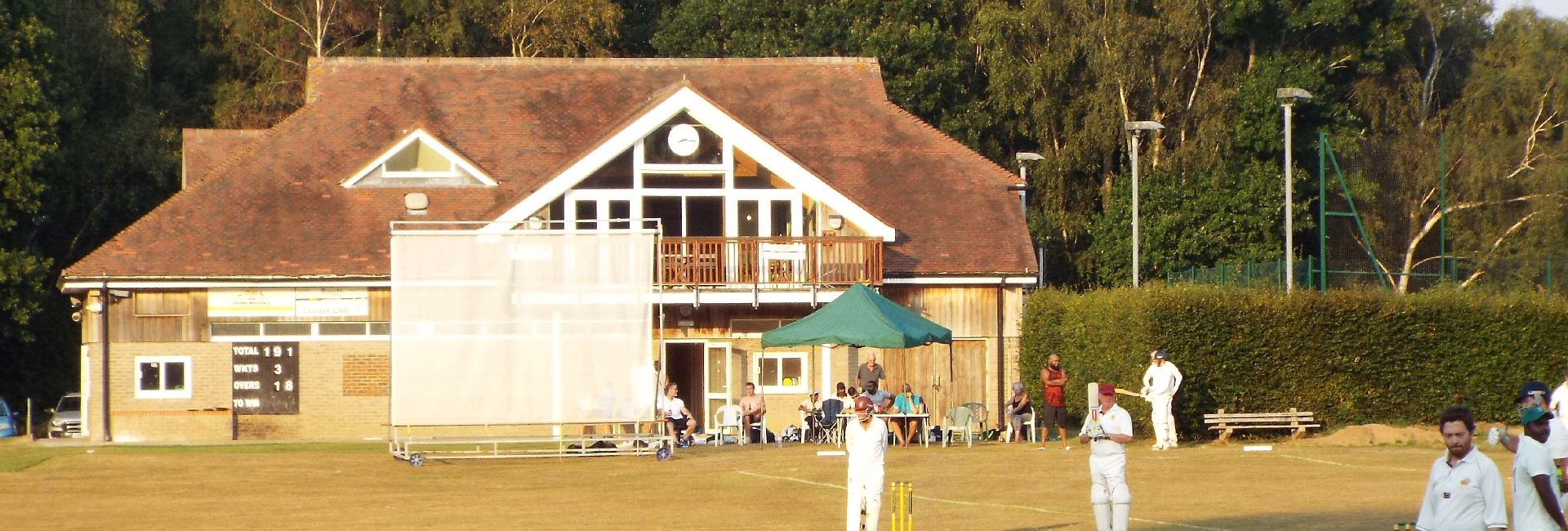Forest Row Cricket Club's pavilion in evening light during a match, scoreboard reading 191 for 3, with players out on the pitch